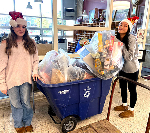 Two women in Santa hats stand beside a blue rolling bin filled with bagged toy donations inside a school or community building..