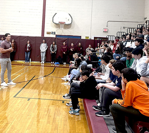 A man stands and speaks to a large group of seated students in a school gymnasium, while staff members watch from the back.