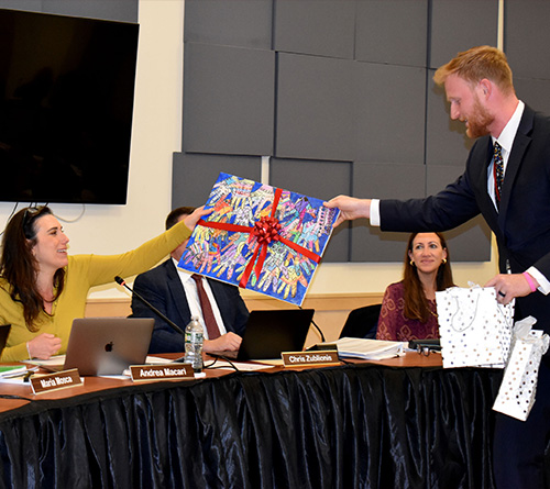A man presents a colorful gift to a smiling woman during a board meeting.