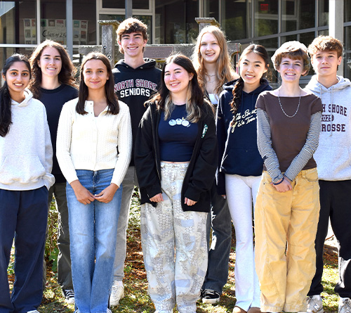A group of smiling high school students stands together outdoors in front of a school building on a sunny day.