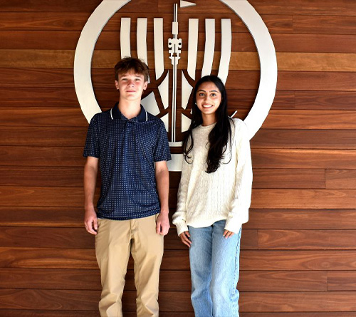 Two students standing in front of a school emblem on a wooden wall