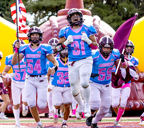 Football players from the Shore team run onto the field through an inflatable tunnel, waving flags and showing excitement before the game.