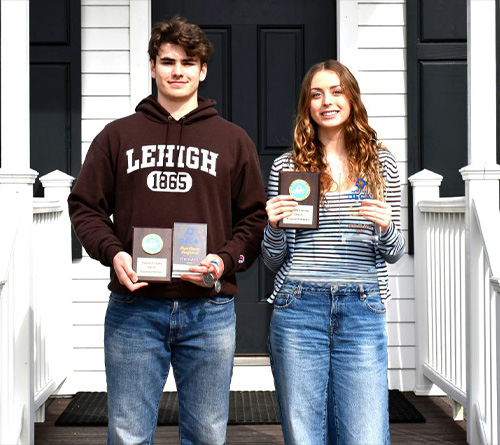Two students standing on a front porch holding DECA awards and medals, smiling after a competition achievement.