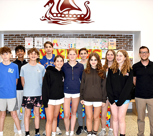 Group of students and a teacher standing together indoors, posing in front of a wall with colorful posters and a Viking ship logo.