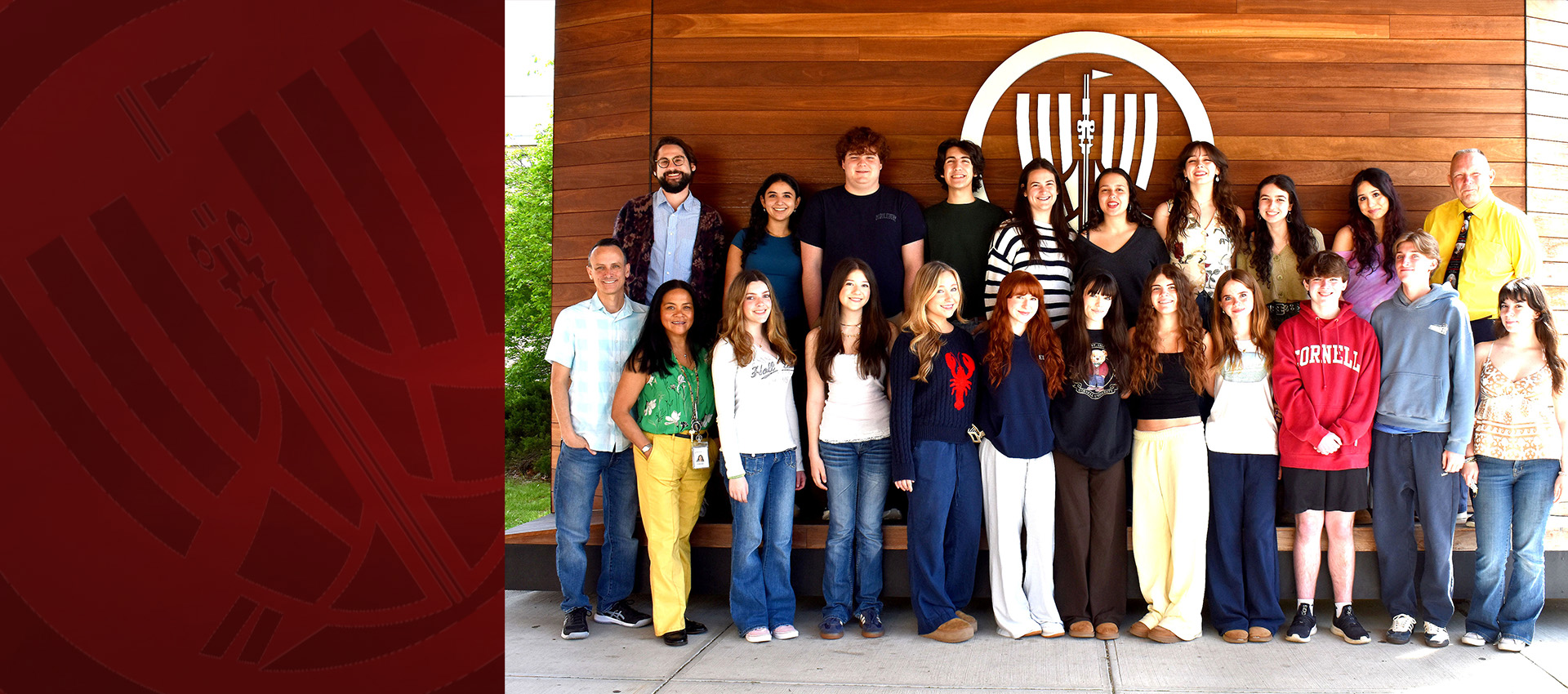 A group of students and a few adults standing together in two rows in front of a wooden wall with a circular emblem, smiling at the camera.