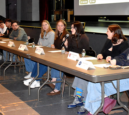 Students seated at a panel discussion on stage, one speaking into a microphone.