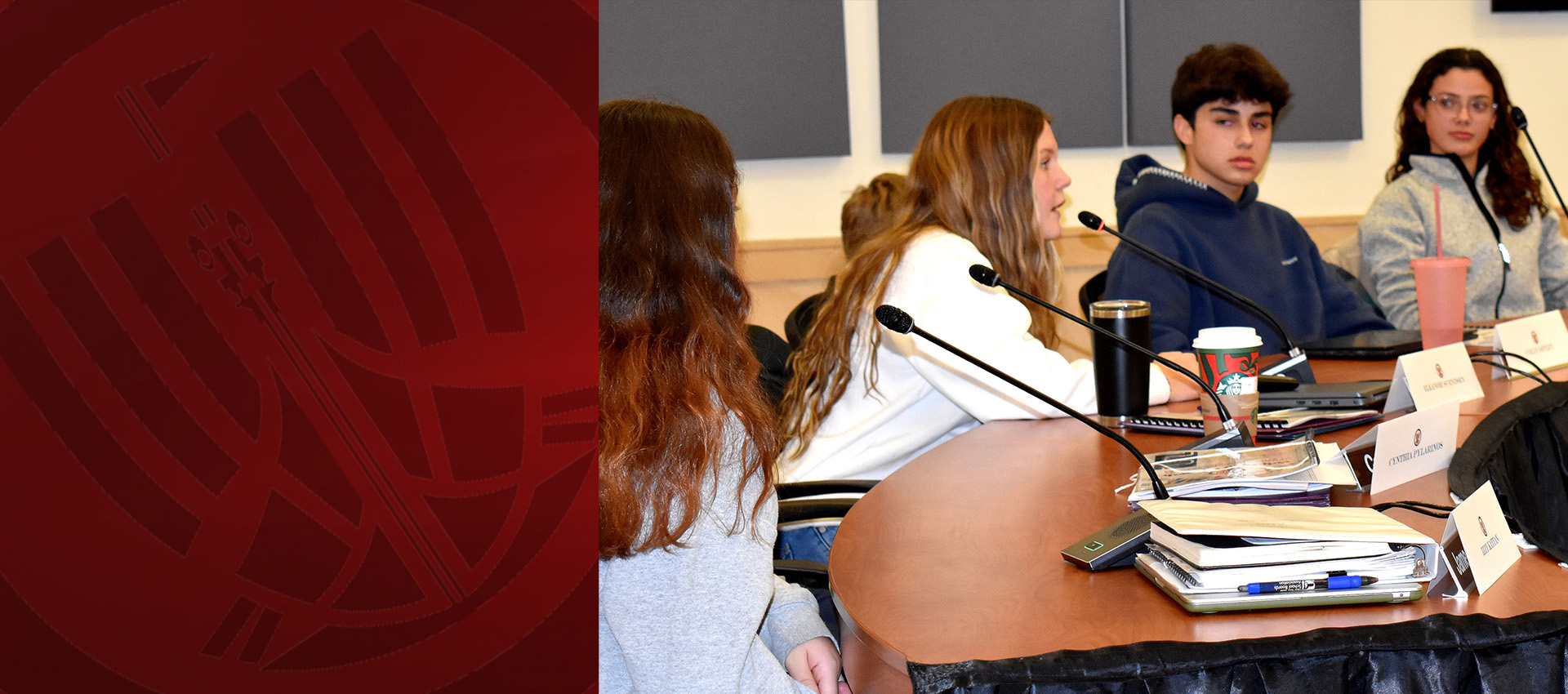 Students seated around a conference table speaking into microphones during a classroom discussion.
