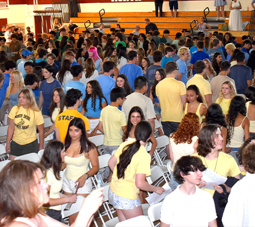 A large group of high school students gathered in a gymnasium, standing between rows of white chairs during a school event.