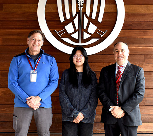 Three people standing and smiling in front of a wooden wall with a large school emblem behind them.