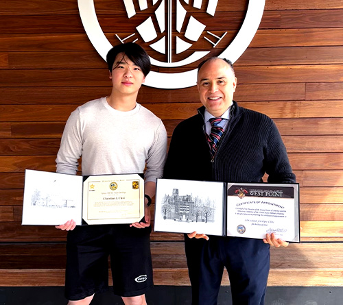 Two people standing in front of a wooden wall with a large emblem, holding framed certificates and awards.