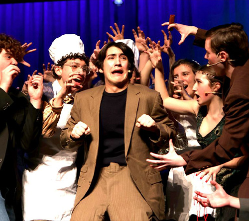 A frightened young man in a suit is surrounded by performers reaching toward him on a brightly lit stage.