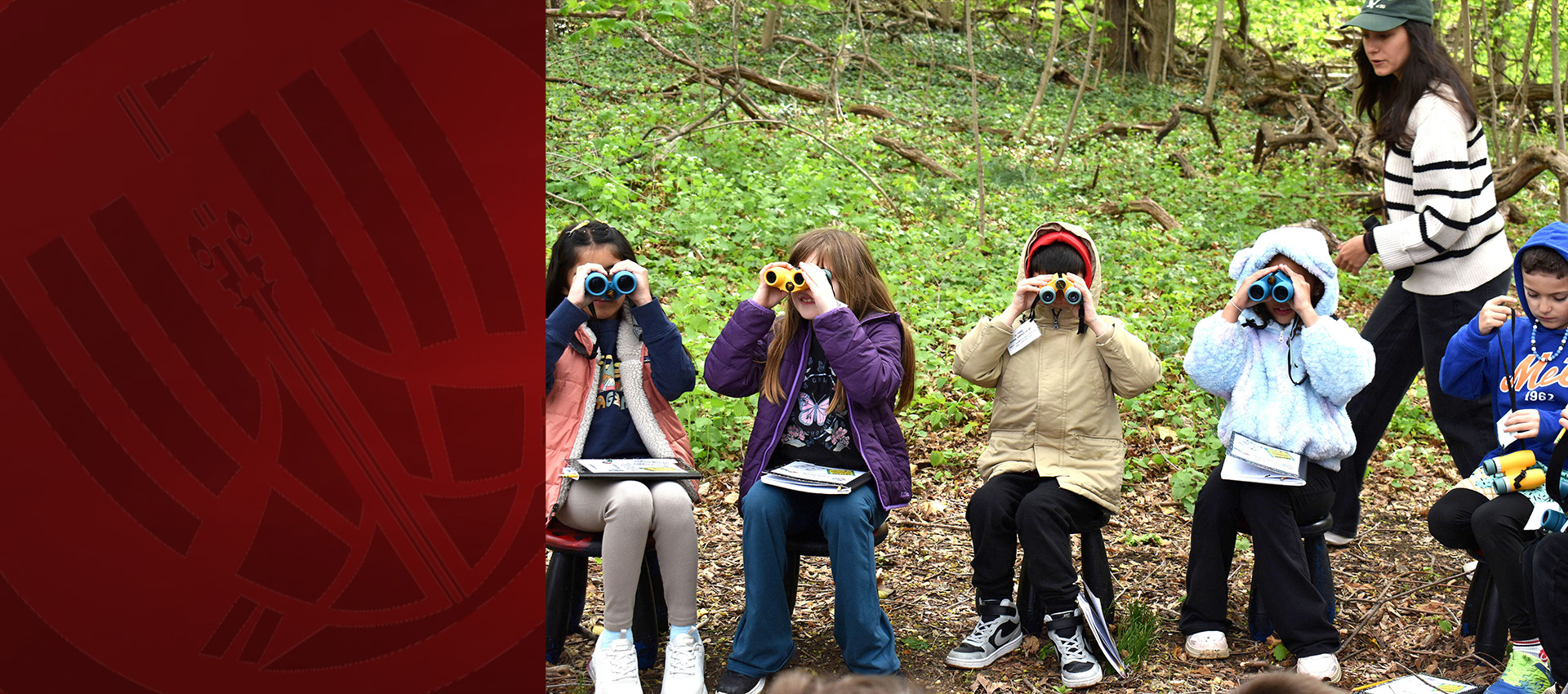 Children sitting outdoors in a forest, using binoculars while a teacher guides them during a nature activity.
