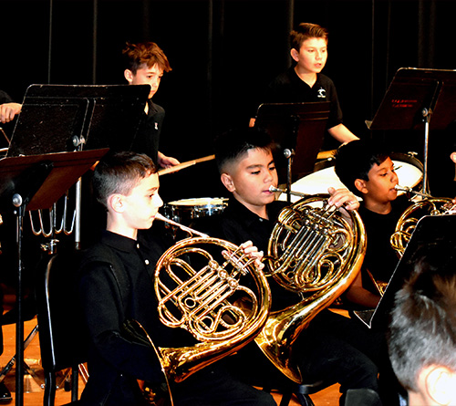 Middle school band students perform on stage during a spring concert, playing French horns and percussion instruments while dressed in black concert attire.