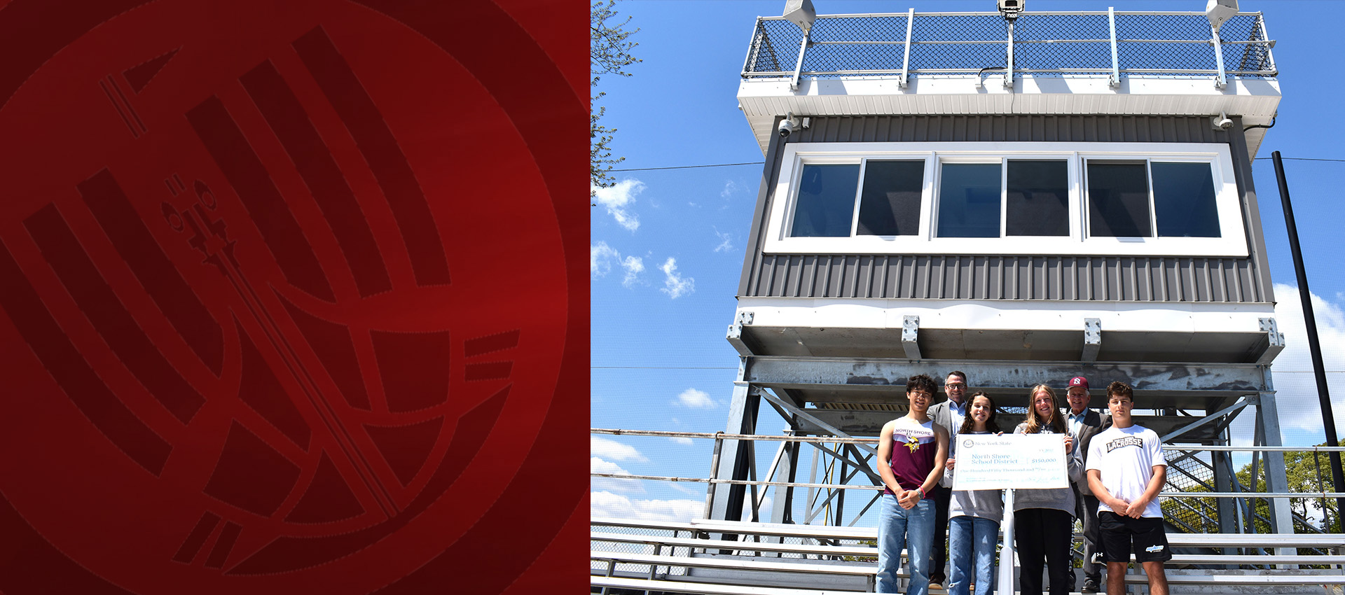 Students and school officials stand in front of a newly constructed high school press box holding a ceremonial grant check at an outdoor athletic field under a clear blue sky.