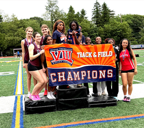Girls track and field team posing with a championship banner and medals on a sports field.