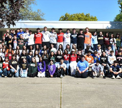 Large group of high school students gathered outside campus wearing college and university shirts.